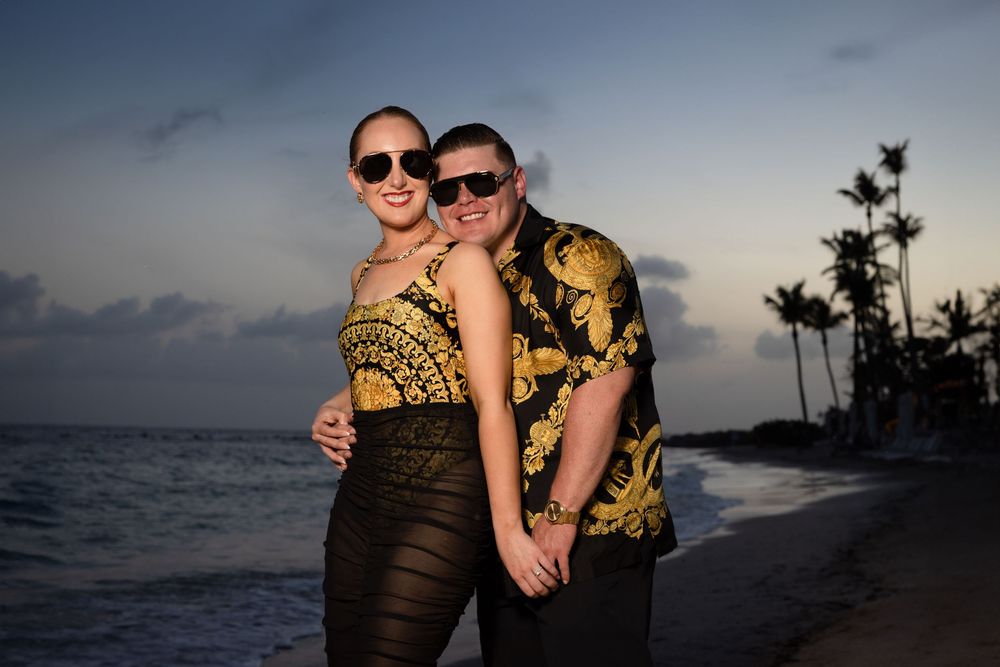 Two people embrace on a tropical beach at sunset with palm trees and ocean waves in the background.
