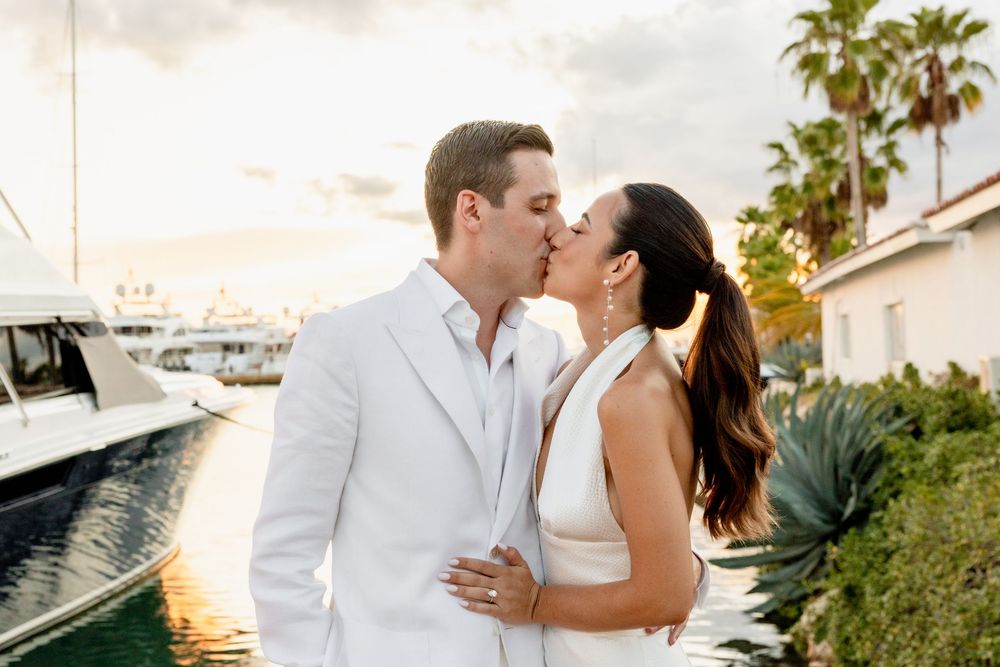 A couple in white outfits share romantic moments on a yacht during sunset with palm trees in the background.