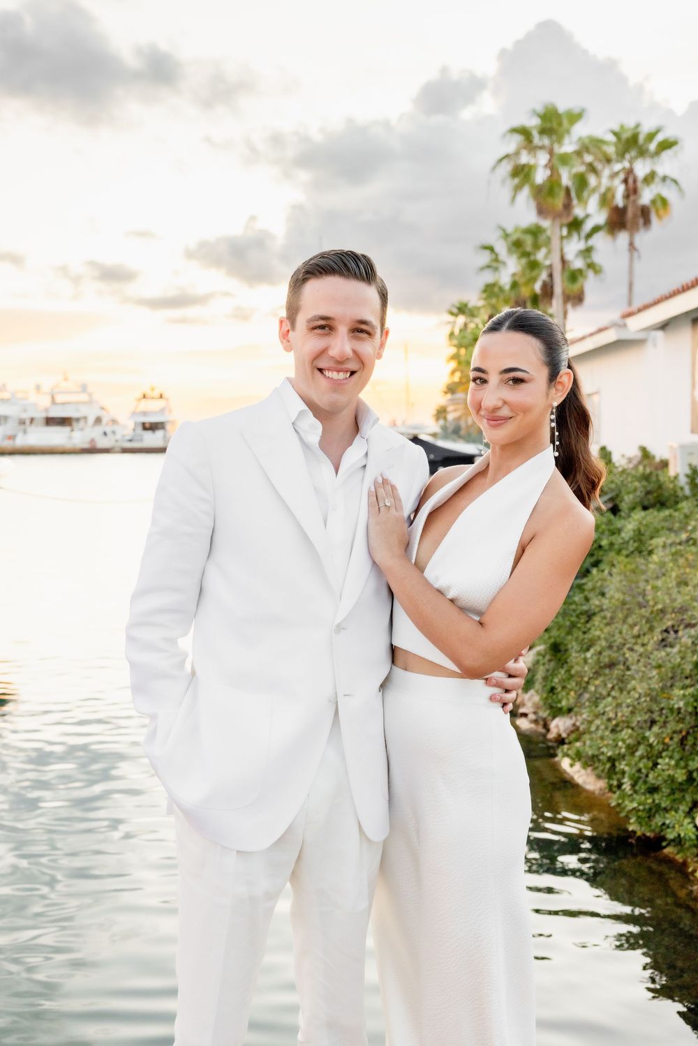A couple in matching white formal attire poses by a marina at sunset with palm trees in the background.