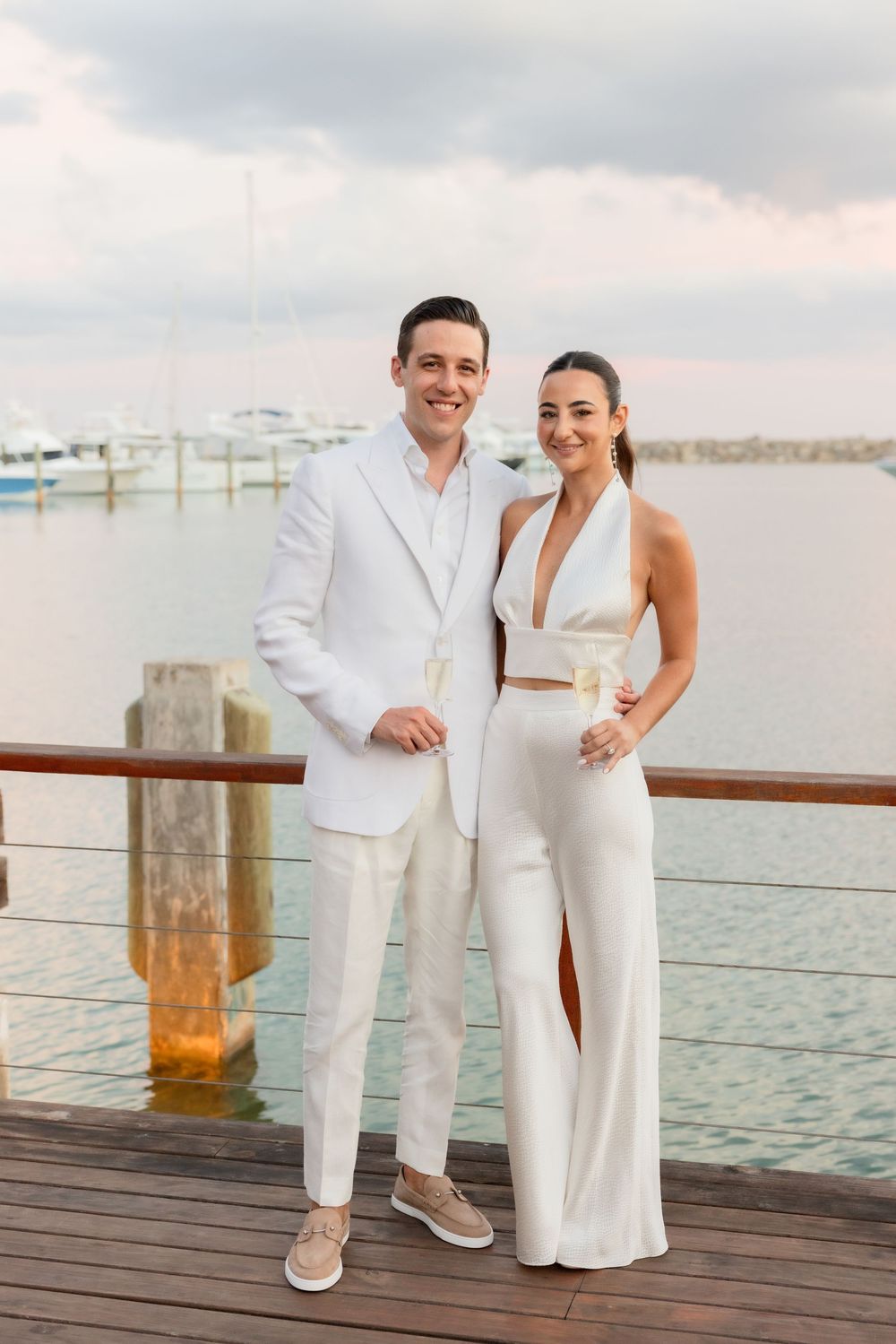 A couple in coordinating white outfits poses together on a wooden dock by the water during sunset.