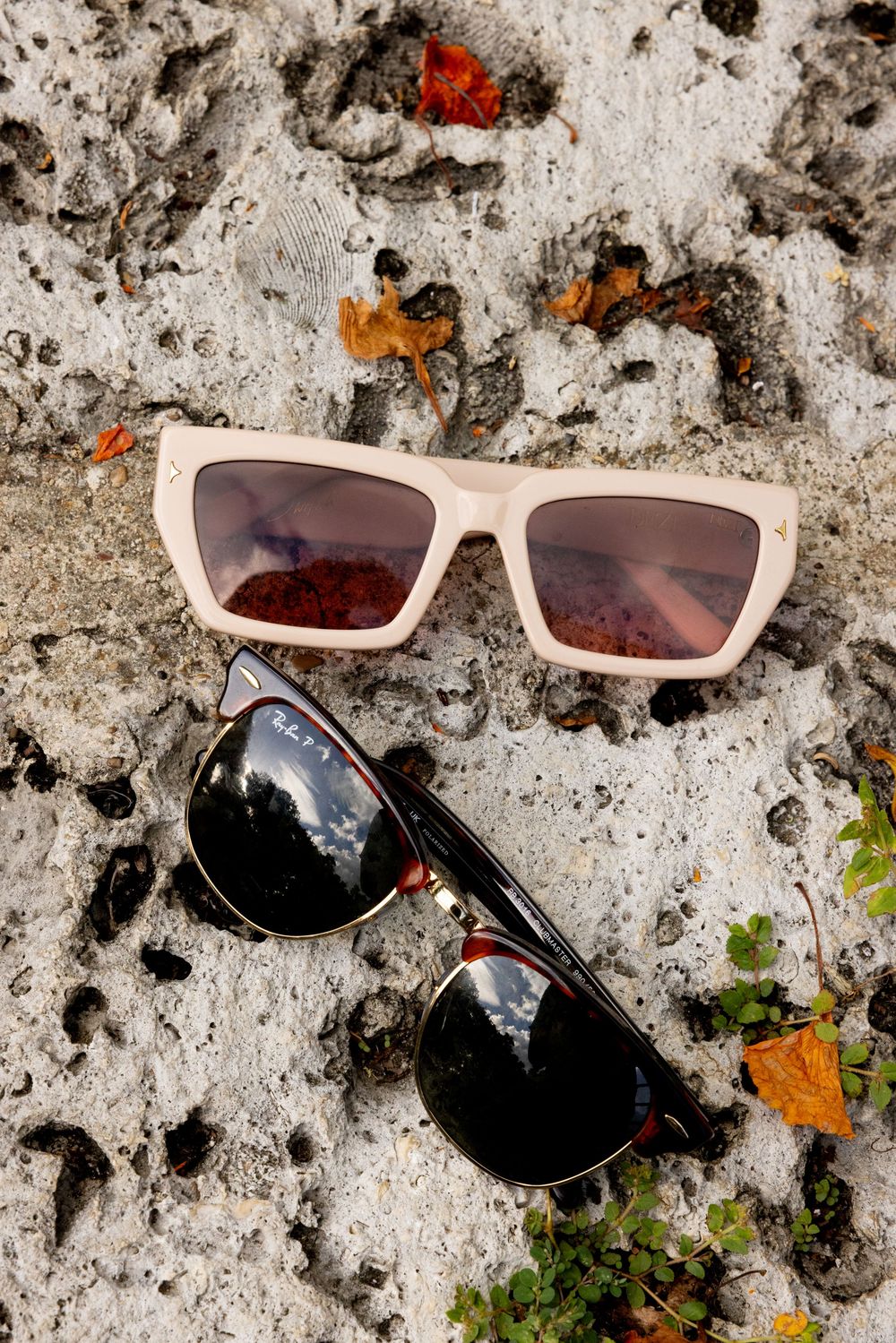 Broken sunglasses with beige frames and dark lenses lying on a textured concrete surface with scattered debris.
