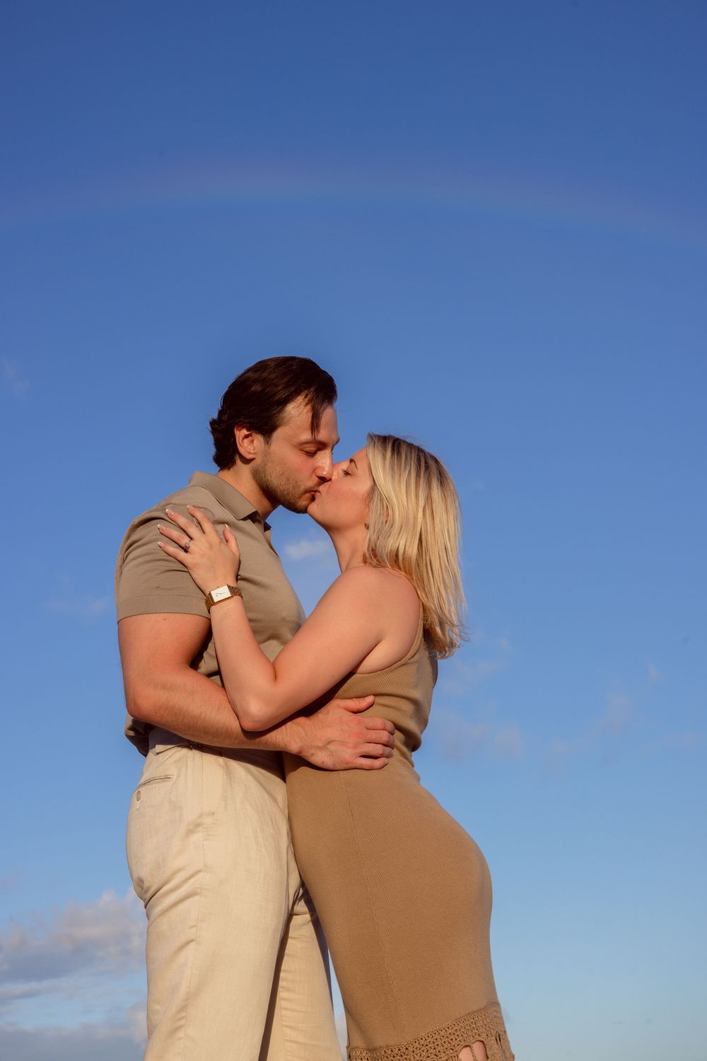 A couple in beige and tan outfits share a romantic kiss against a bright blue sky background.