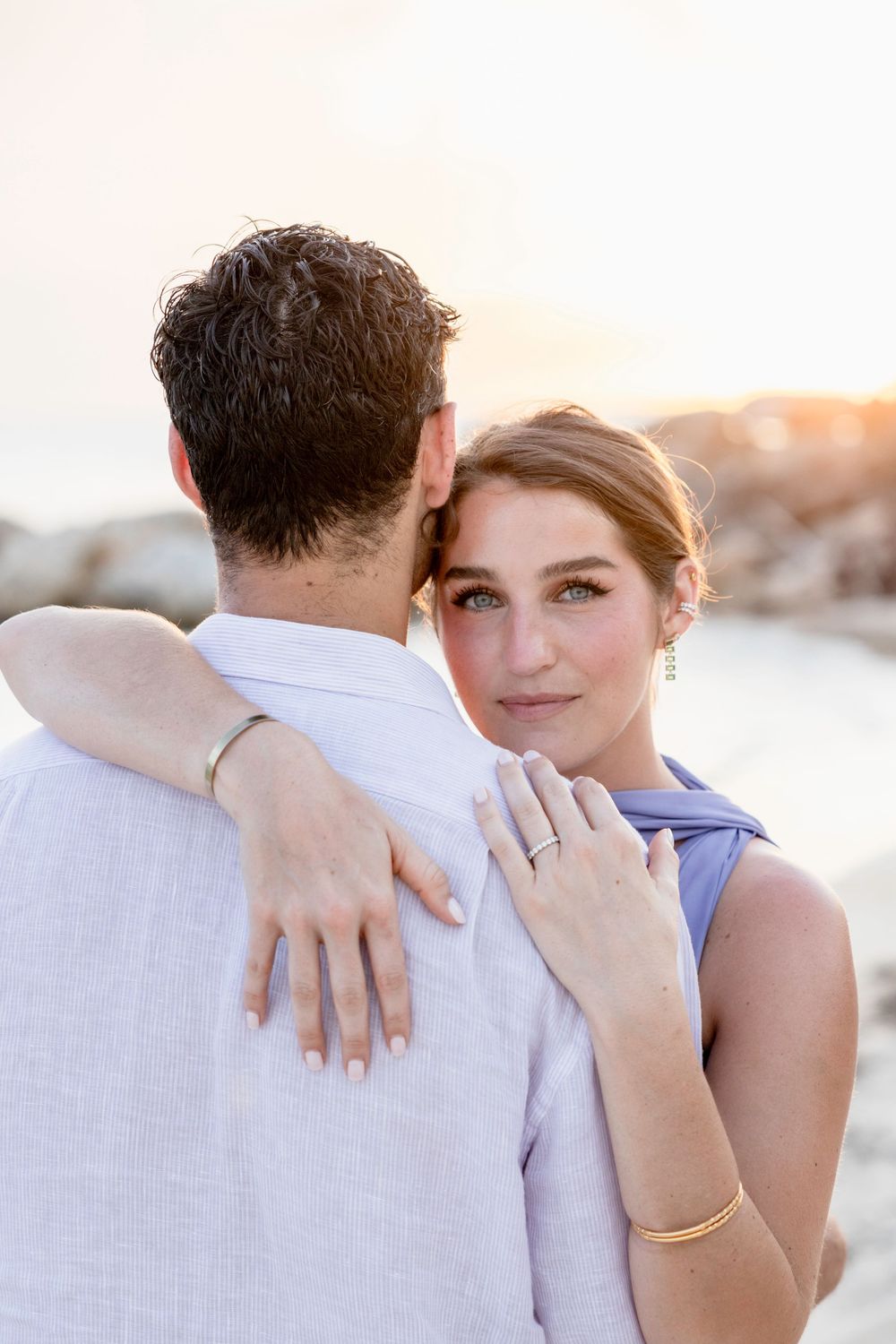 Romantic sunset portrait of a couple embracing on the beach as she shows off her engagement ring.