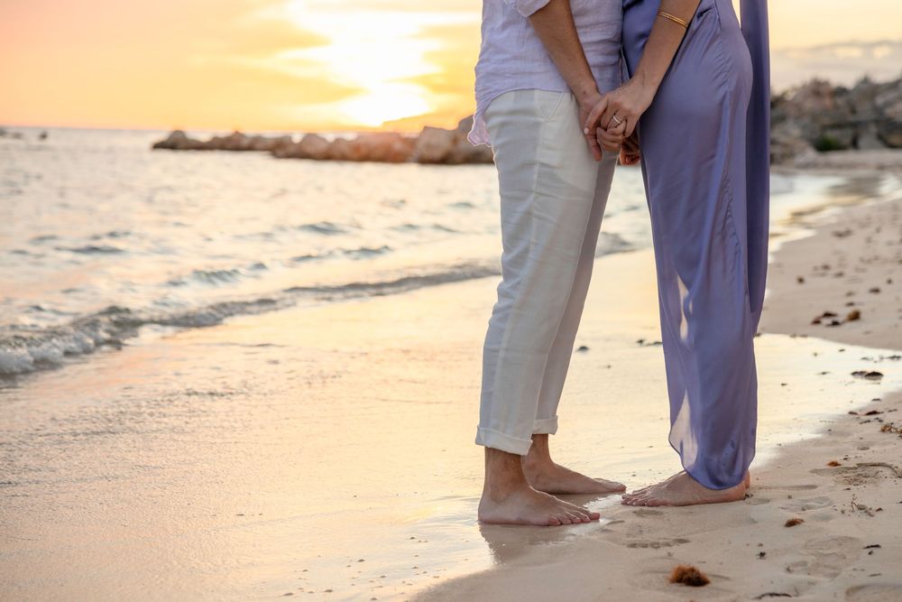 A couple holds hands on a sandy beach at sunset while waves gently roll onto the shore in the background.