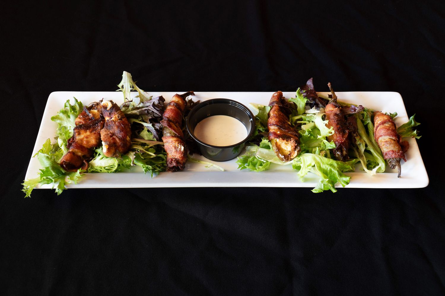 Bacon wrapped scallops served on a white rectangular plate with fresh lettuce and dipping sauce.