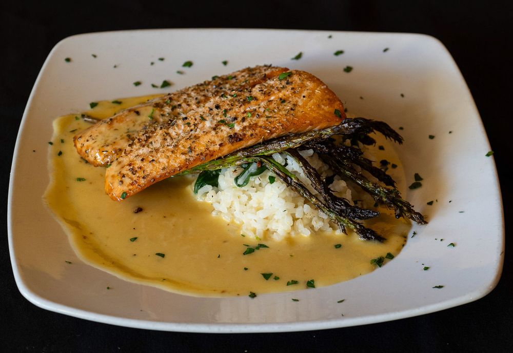 A grilled salmon fillet with sesame seeds served over rice and topped with nori in a creamy sauce on a white plate.