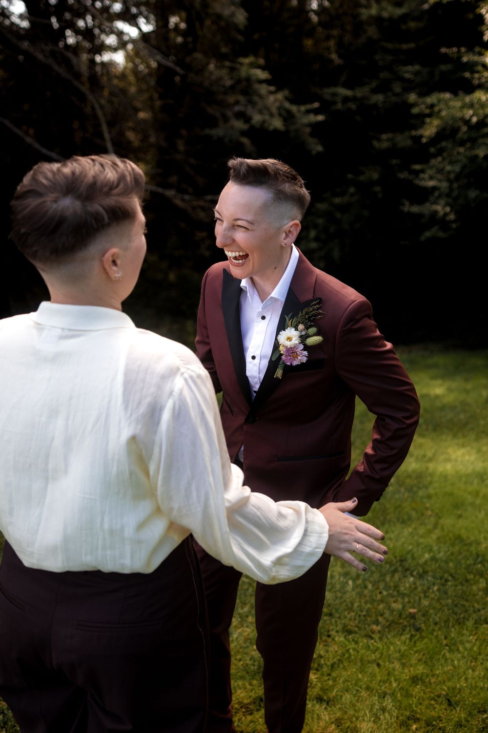 Wedding party members in burgundy and white attire share a candid moment on grassy lawn.
