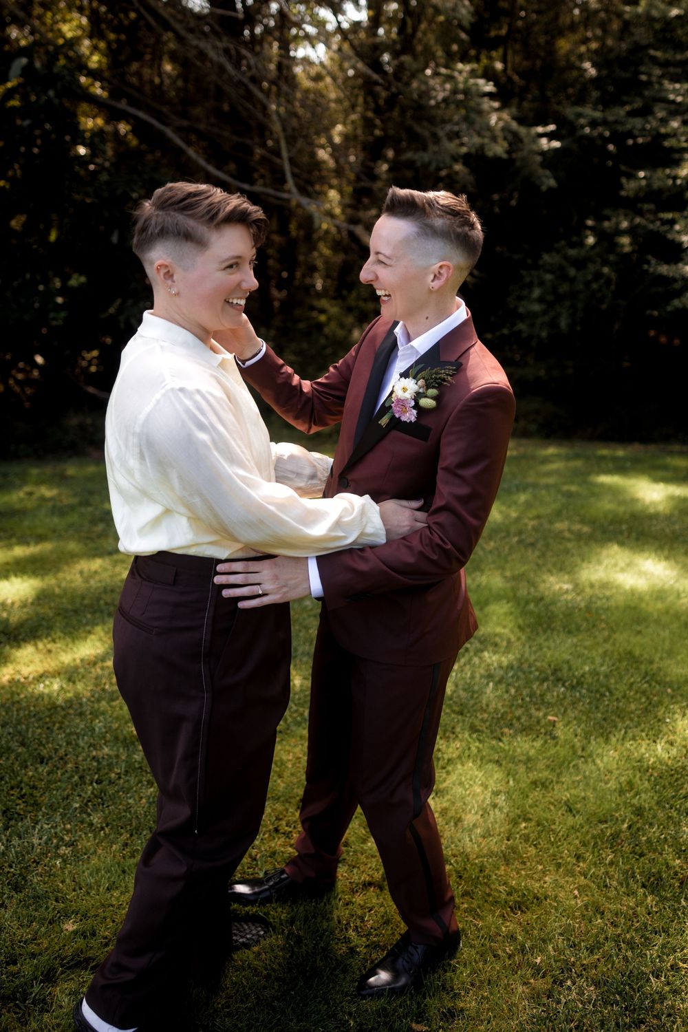 Two people in formal attire sharing playful moments on sunny grass.