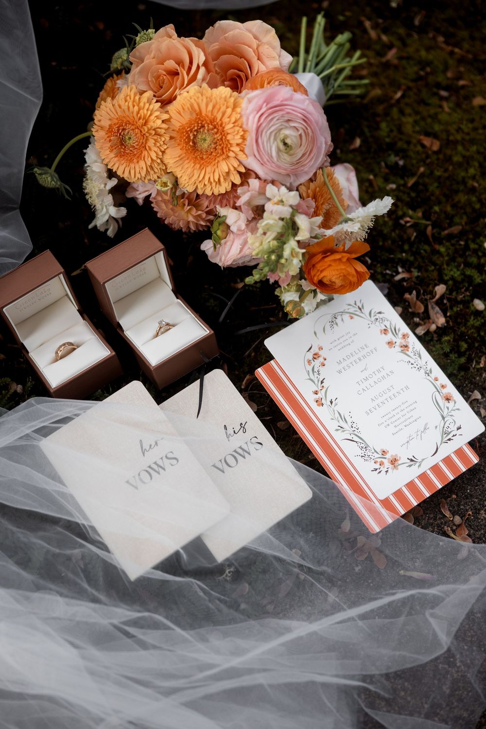 Wedding details including peach and coral floral arrangements, vow books, and ring boxes arranged on white tulle.