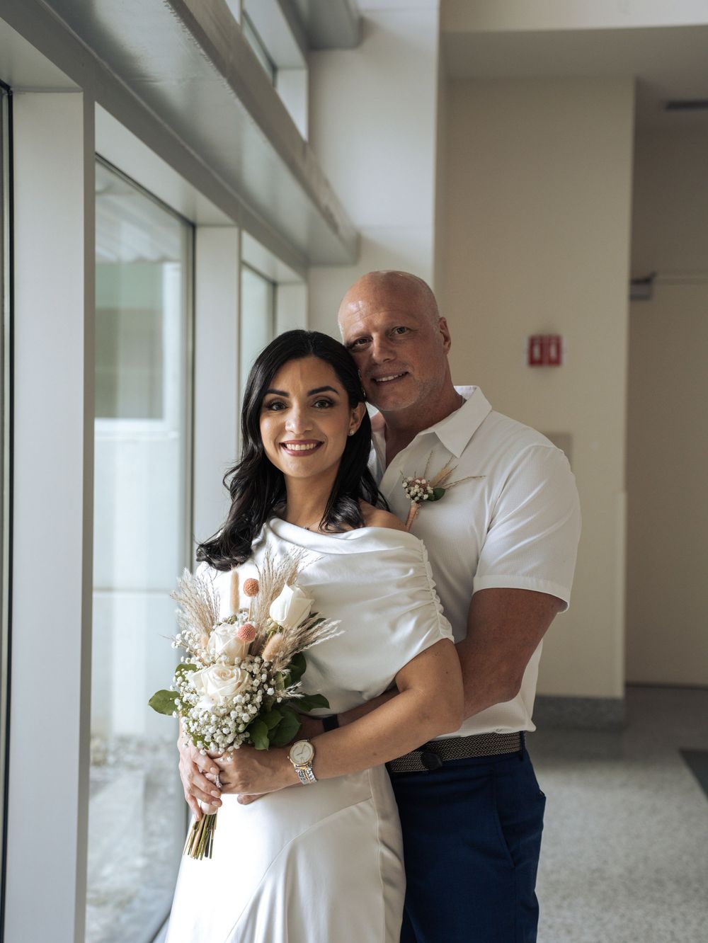 A couple in white outfits embrace near large windows in a bright hallway while holding a small floral bouquet in Naples, Florida