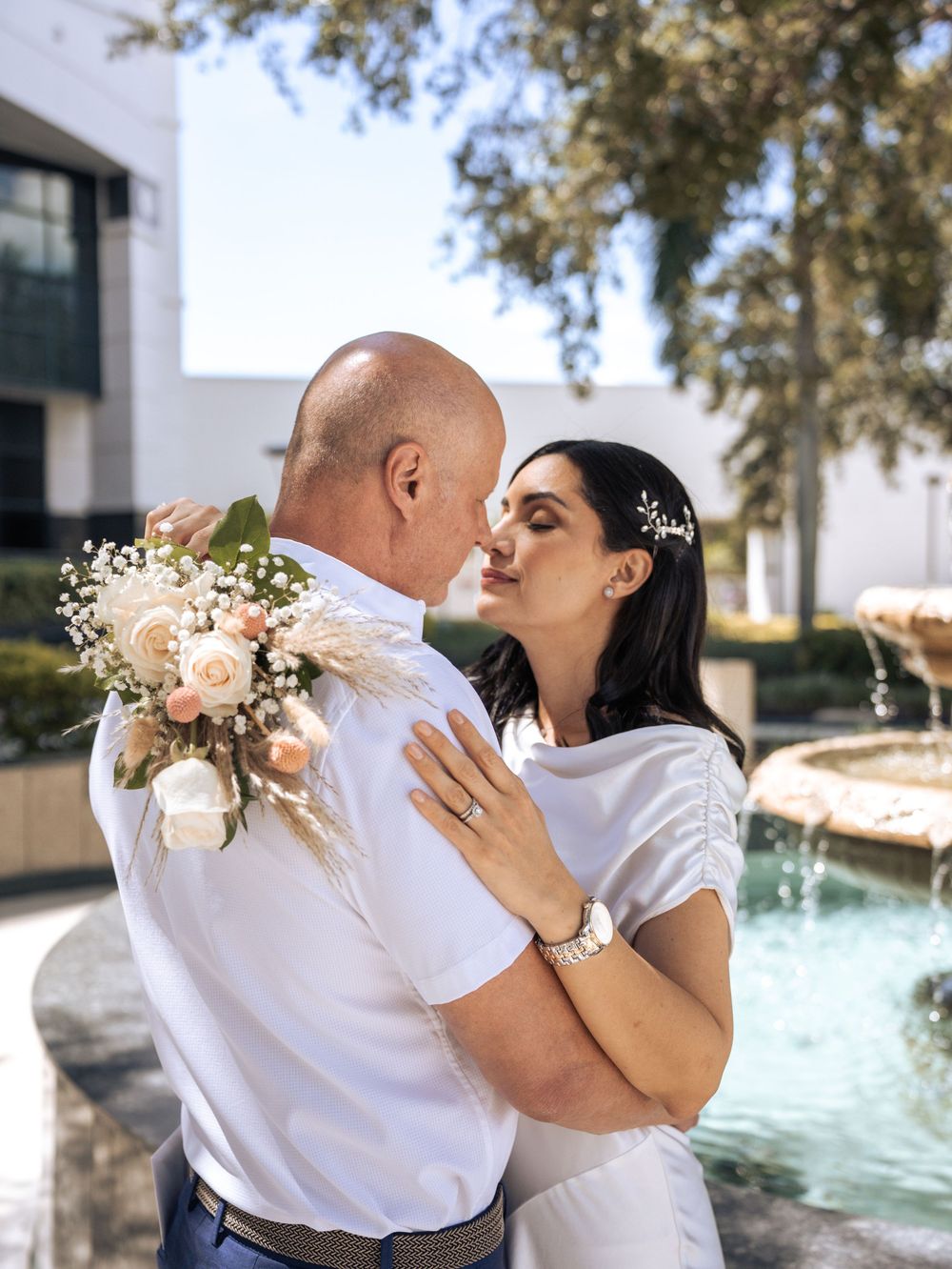 A couple in white outfits shares romantic kisses while dancing by a fountain in a sun-dappled outdoor setting in Naples, Florida