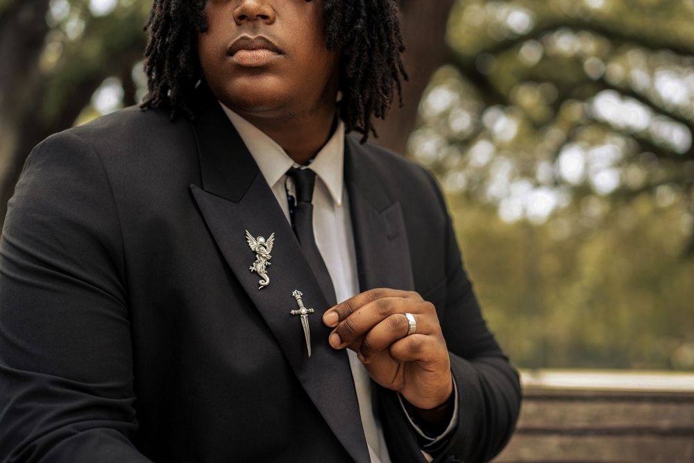 Person wearing a black suit and tie holding a lapel pin against a blurred outdoor background.