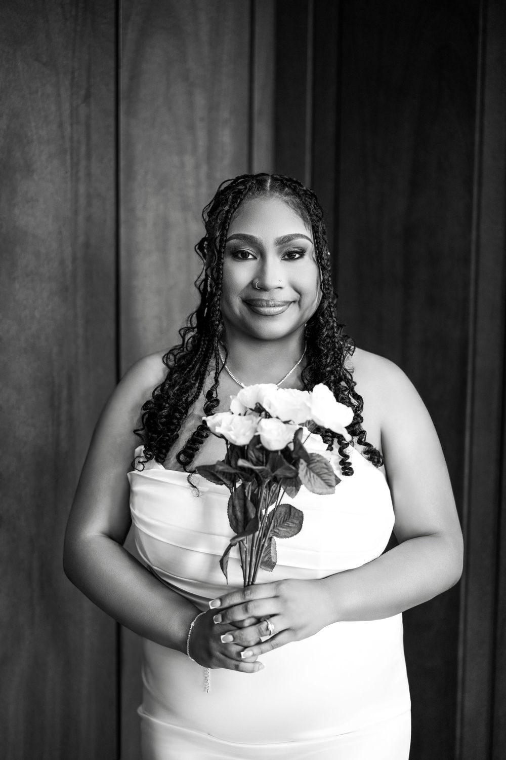 A black and white portrait of a bride in a white dress holding a bouquet of roses against a dark background.