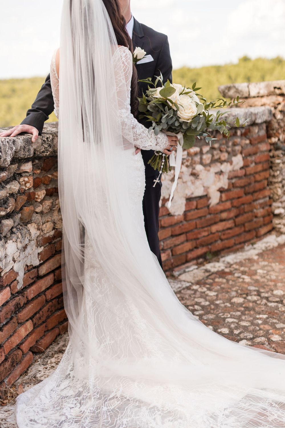 Flowing white wedding dress train and veil draped elegantly against rustic brick wall.