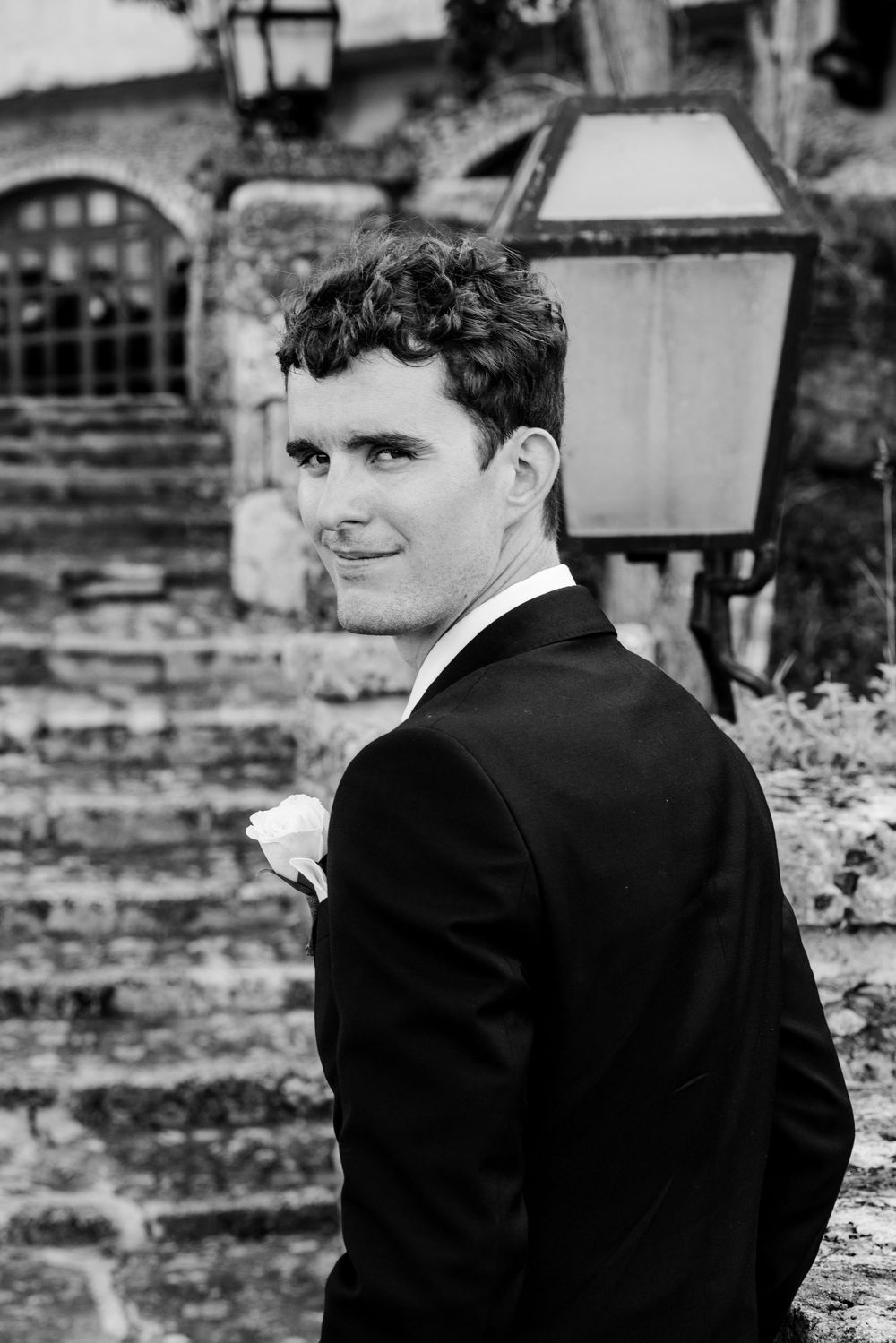 Black and white portrait of a groom in a suit looking over his shoulder with a stone wall and lantern in the background.