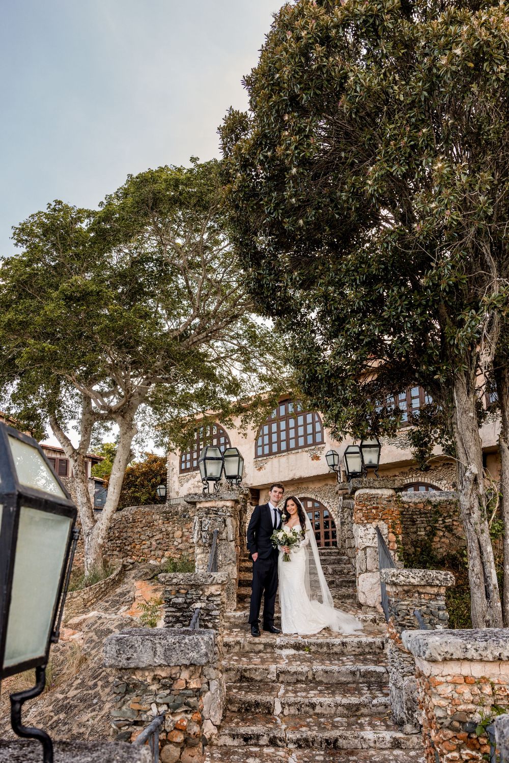 A wedding couple stands on rustic stone steps leading to a historic building surrounded by lush trees.