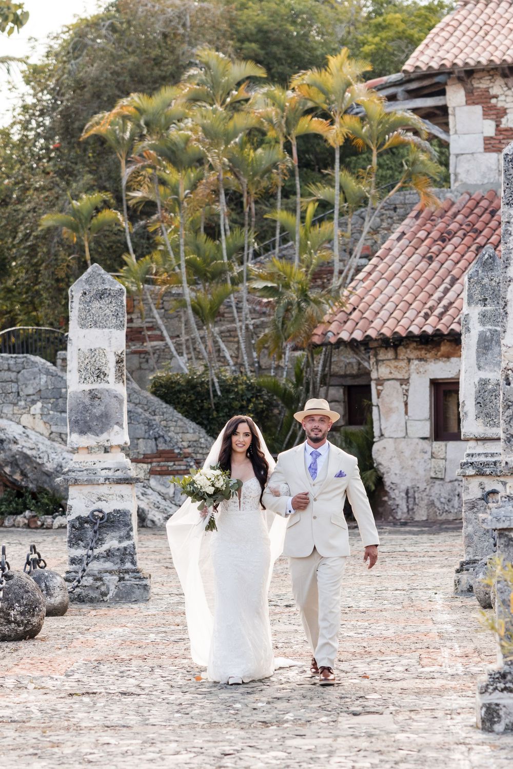 A couple in white wedding attire walks together on a stone courtyard surrounded by palm trees and rustic stone walls.