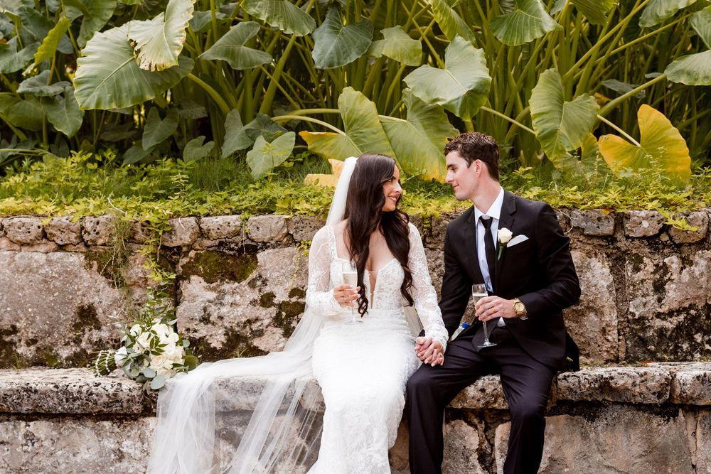 A romantic wedding couple walks together through a garden setting with stone walls and lush greenery.