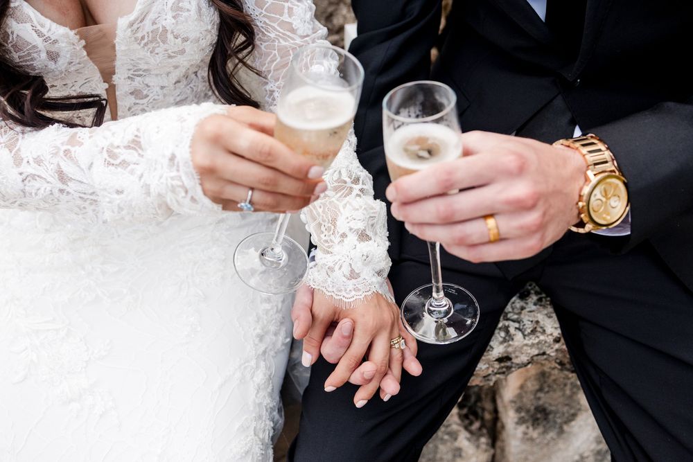 Hands of a bride and groom holding champagne glasses while wearing formal wedding attire.