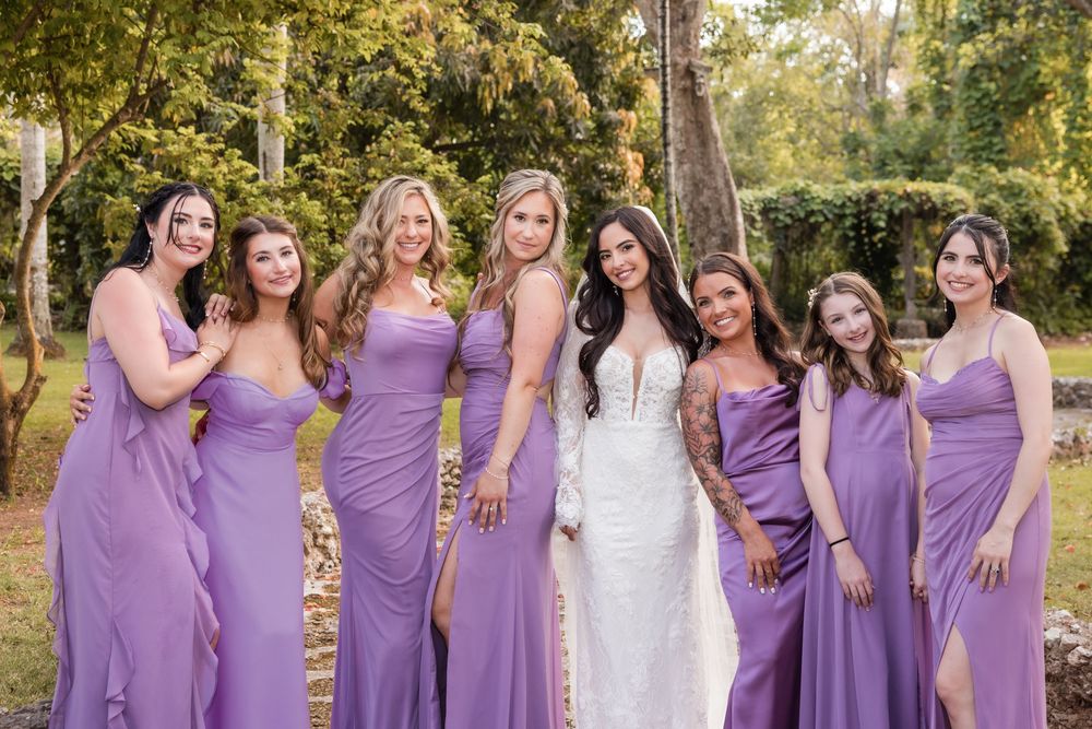 Bridesmaids in matching purple dresses stand together for a group photo in a garden setting.