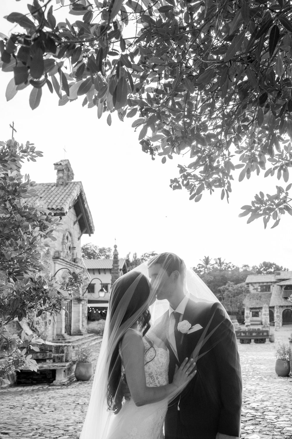 A romantic couple shares a tender moment under tree branches in a garden setting.