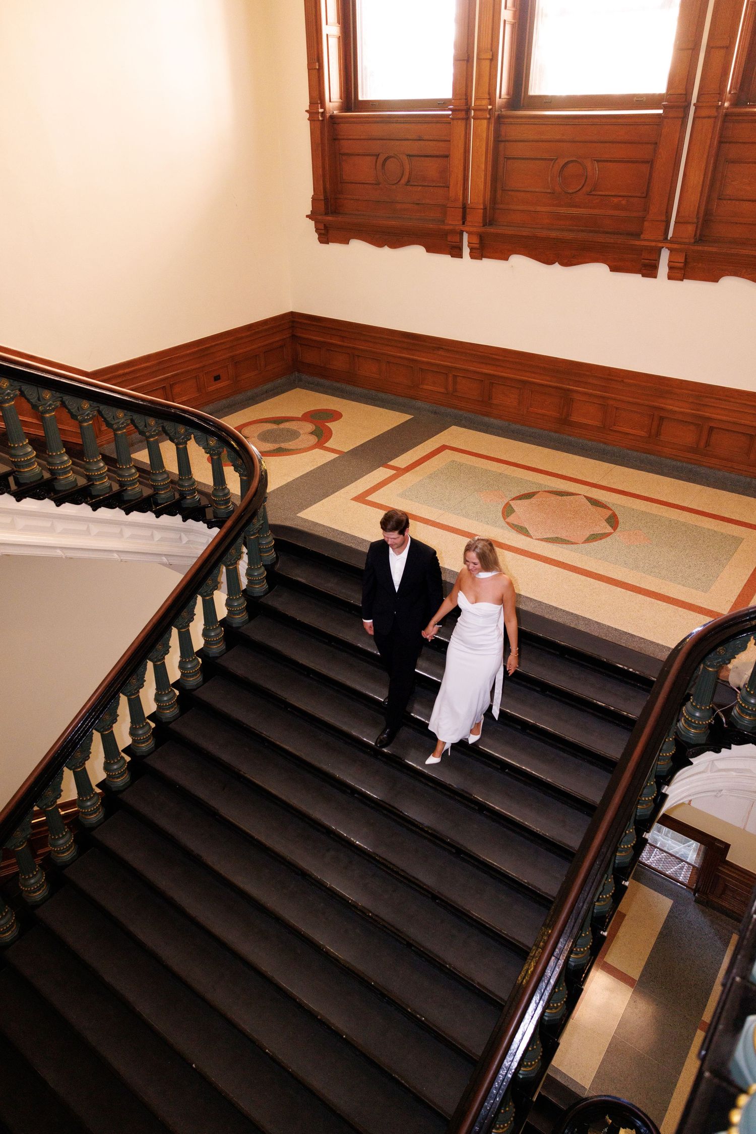 Aerial view of couple descending grand wooden staircase in formal attire.