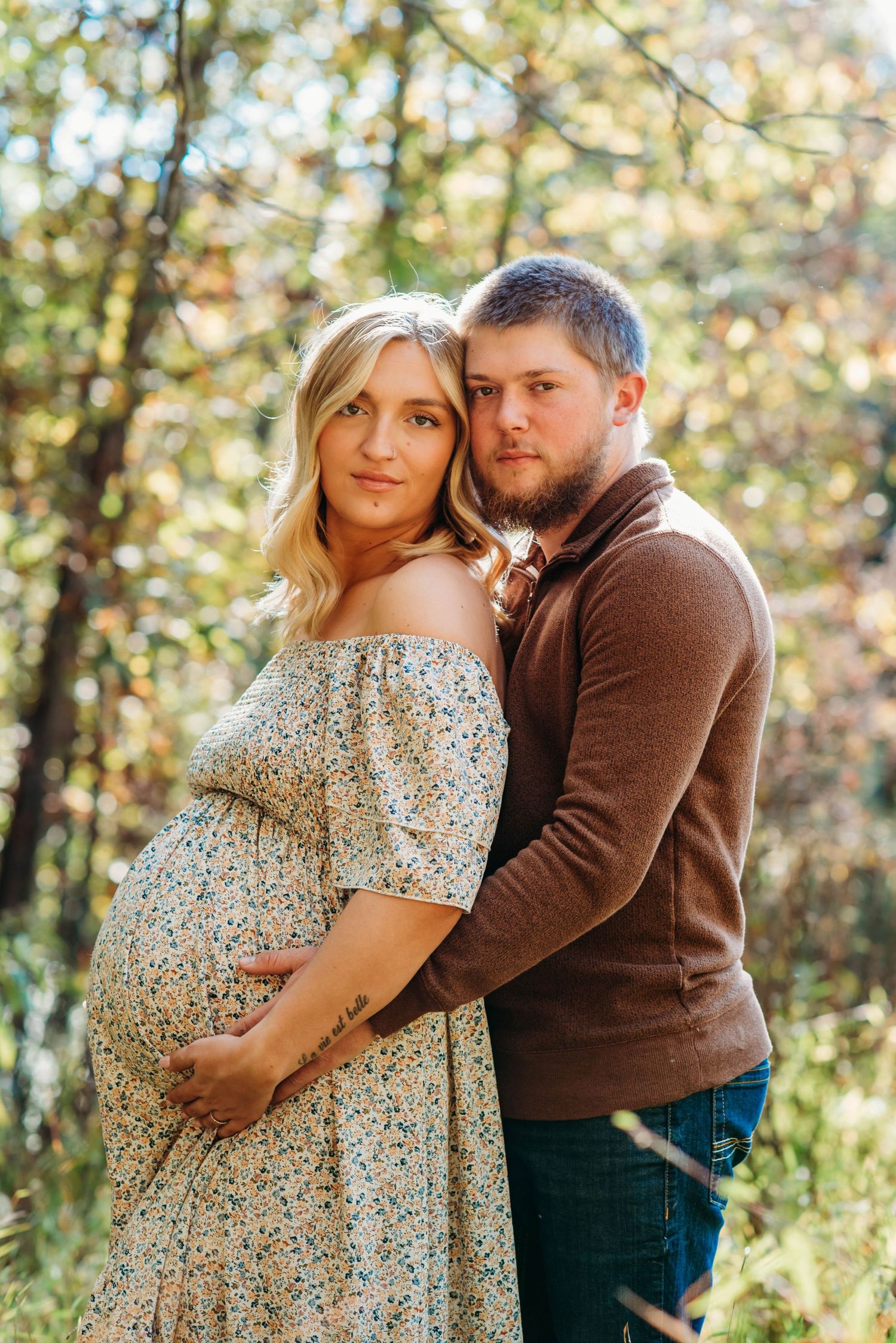 A couple embraces in a sun-dappled forest setting wearing a floral dress and brown sweater during an autumn photoshoot.