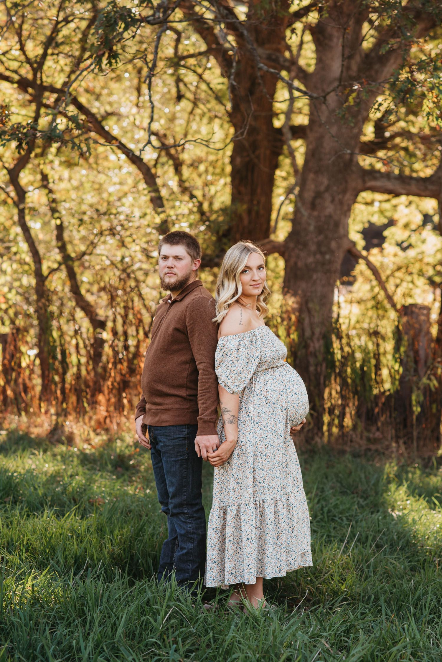 A couple stands together in a grassy field with golden sunlit trees in the background during a sunset photoshoot.