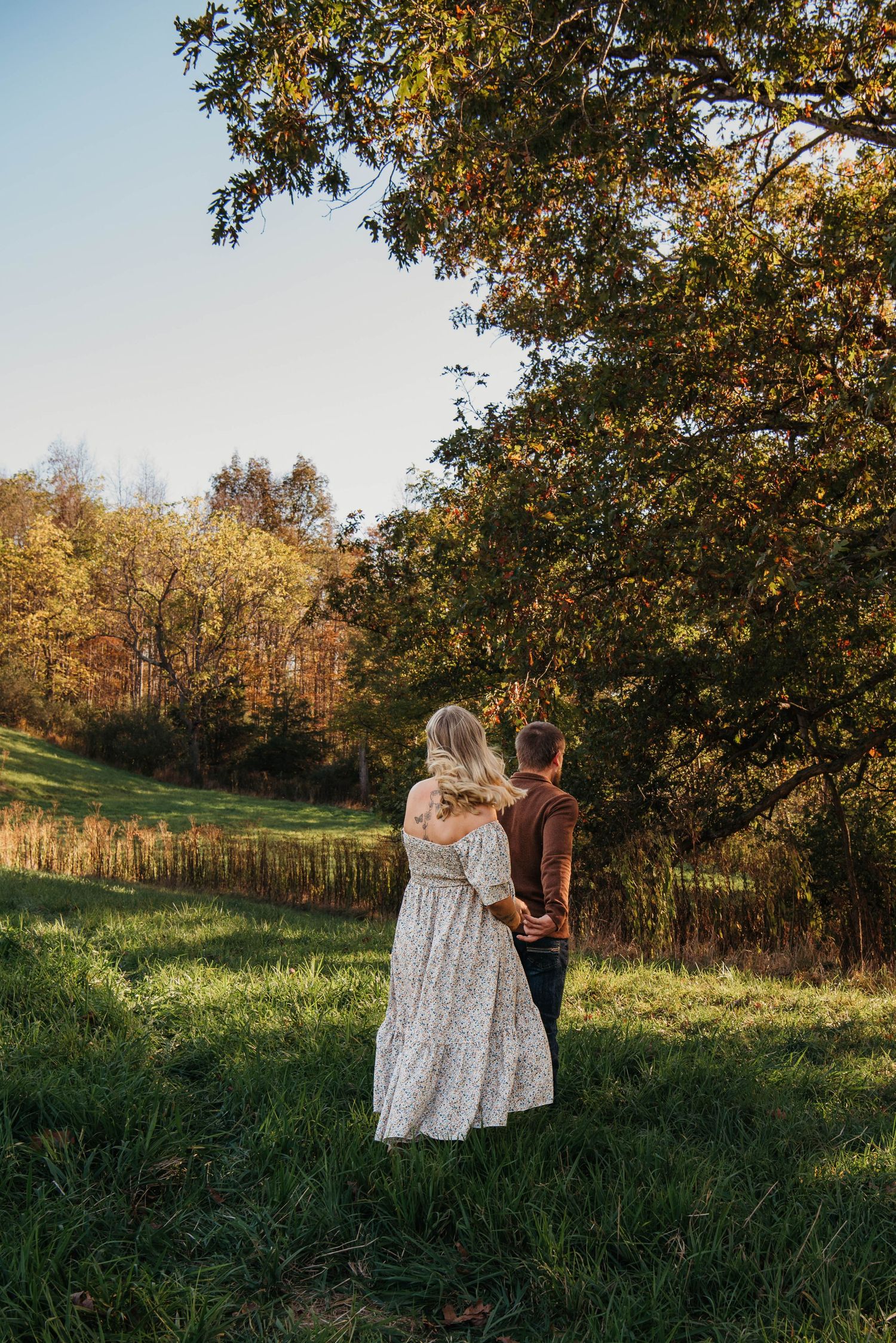 A couple in matching outfits walks through a sunlit autumn meadow surrounded by golden trees at sunset.