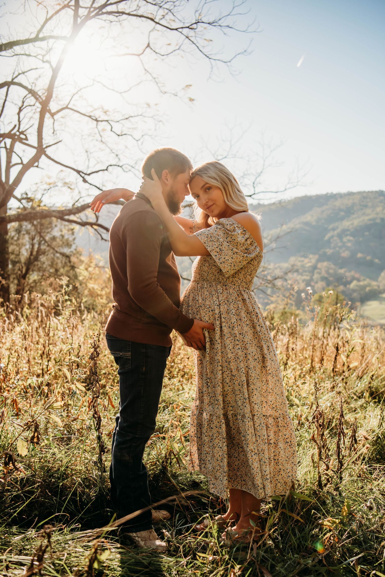 A couple dances together in a sunlit field surrounded by tall grass and mountains in the background.