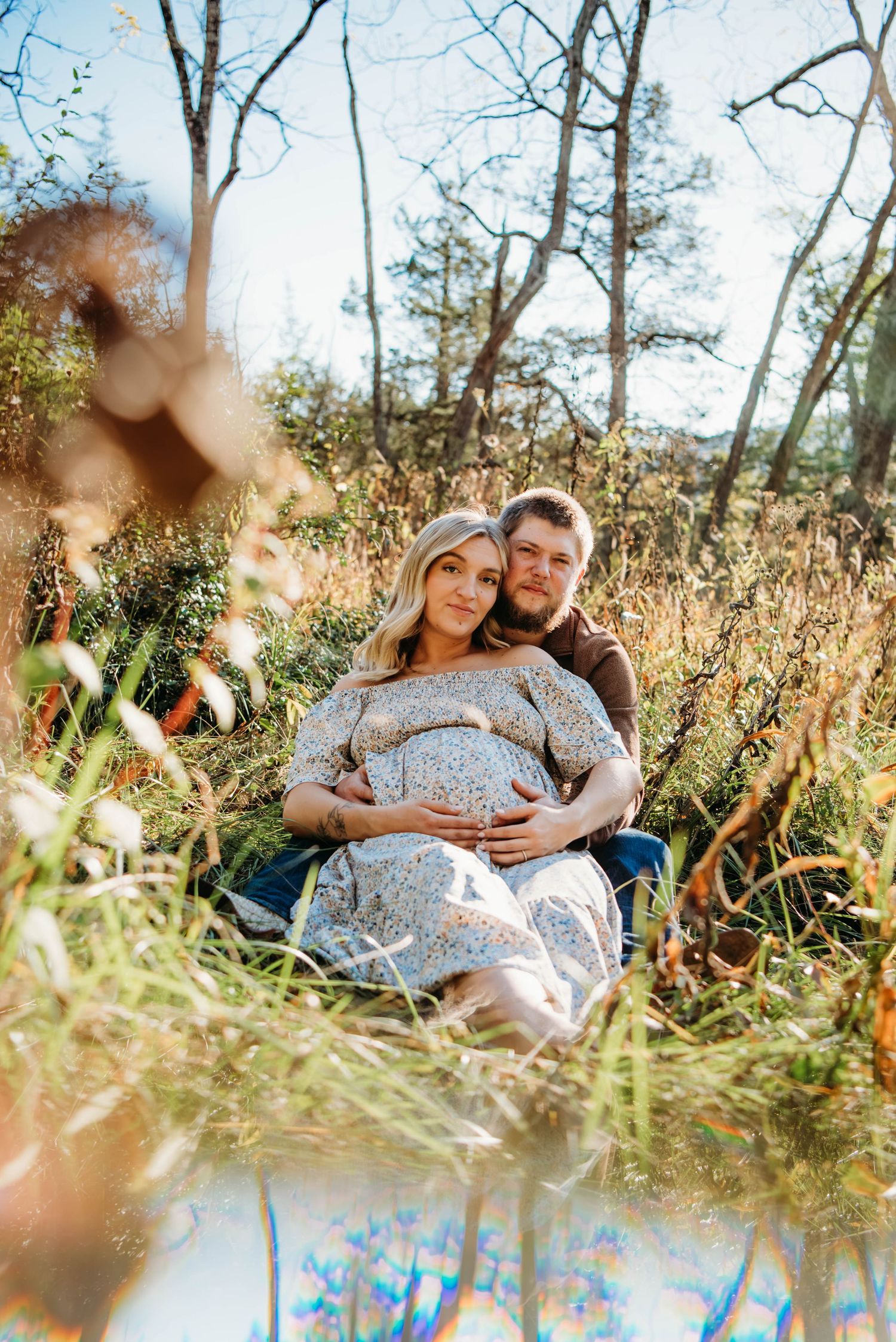 A couple sits together in tall grass during a sunny autumn maternity photoshoot near a reflective pond.