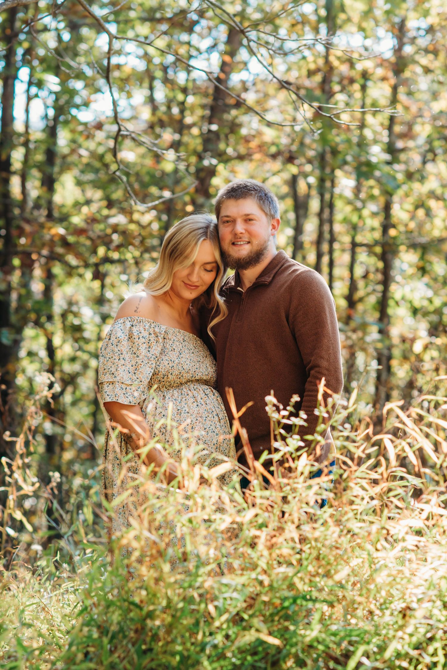 A romantic couple embraces in a sunlit forest with golden grasses and pine trees in the background.