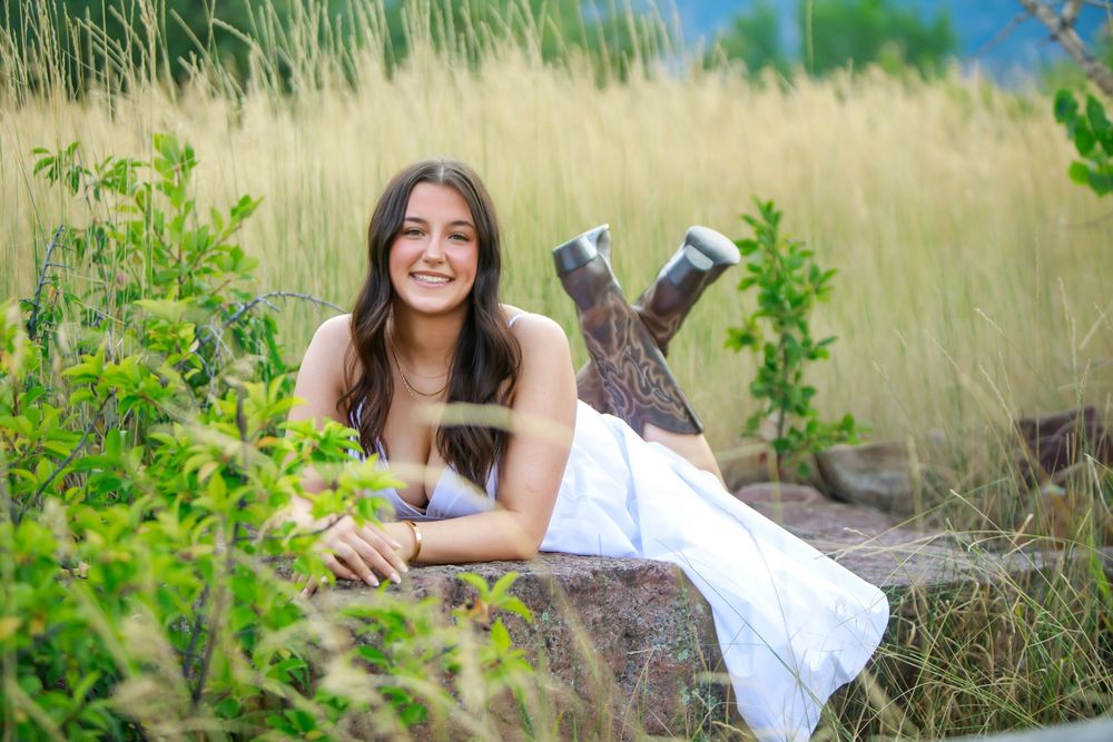 A person in a white dress reclines on grass in a natural outdoor setting during a photo session.