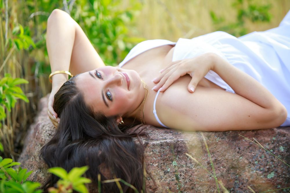 Person with long dark hair lying on a rock outdoors in a white dress, surrounded by green foliage.