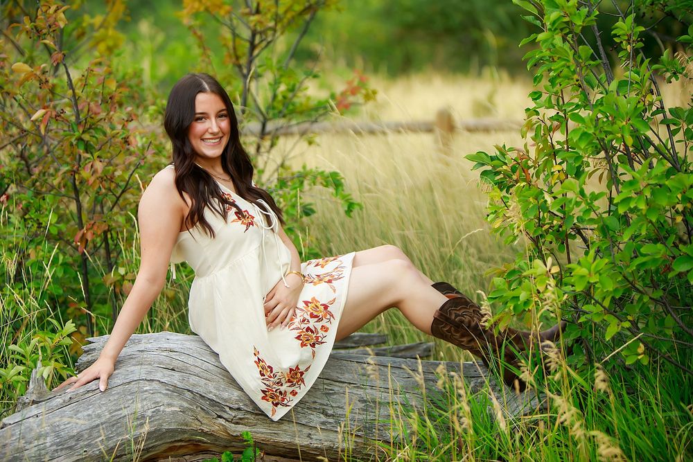 A person in a white floral dress sits casually on a large rock surrounded by lush green foliage in a natural setting.