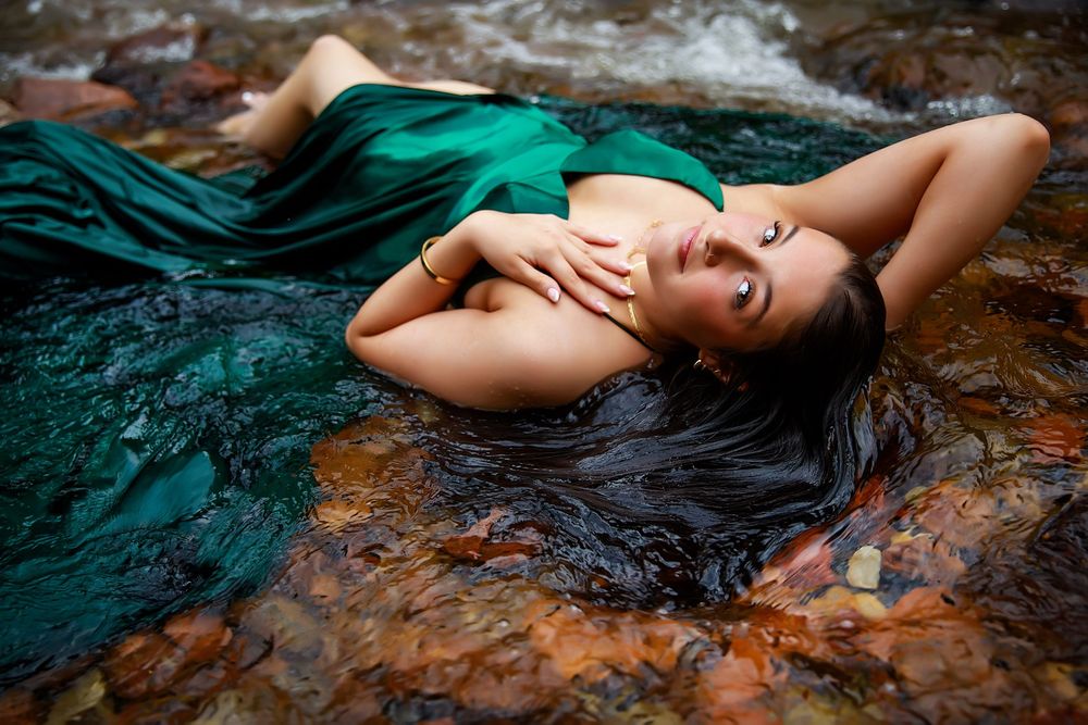A figure in a teal dress lies gracefully in shallow flowing water against rocky terrain, creating a dreamy atmosphere.