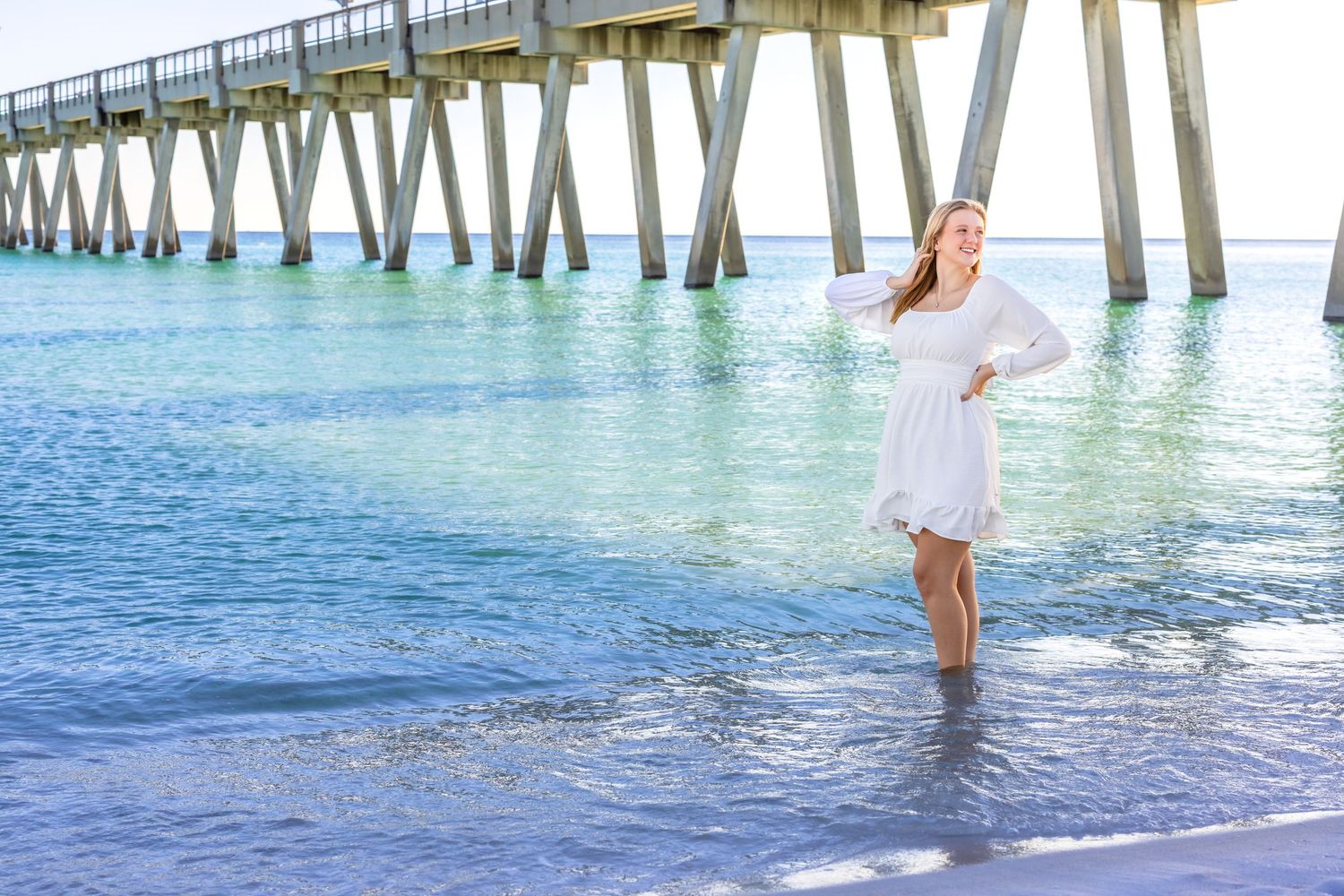 A person in a white dress stands in turquoise ocean water beneath a wooden pier on a sunny beach day.