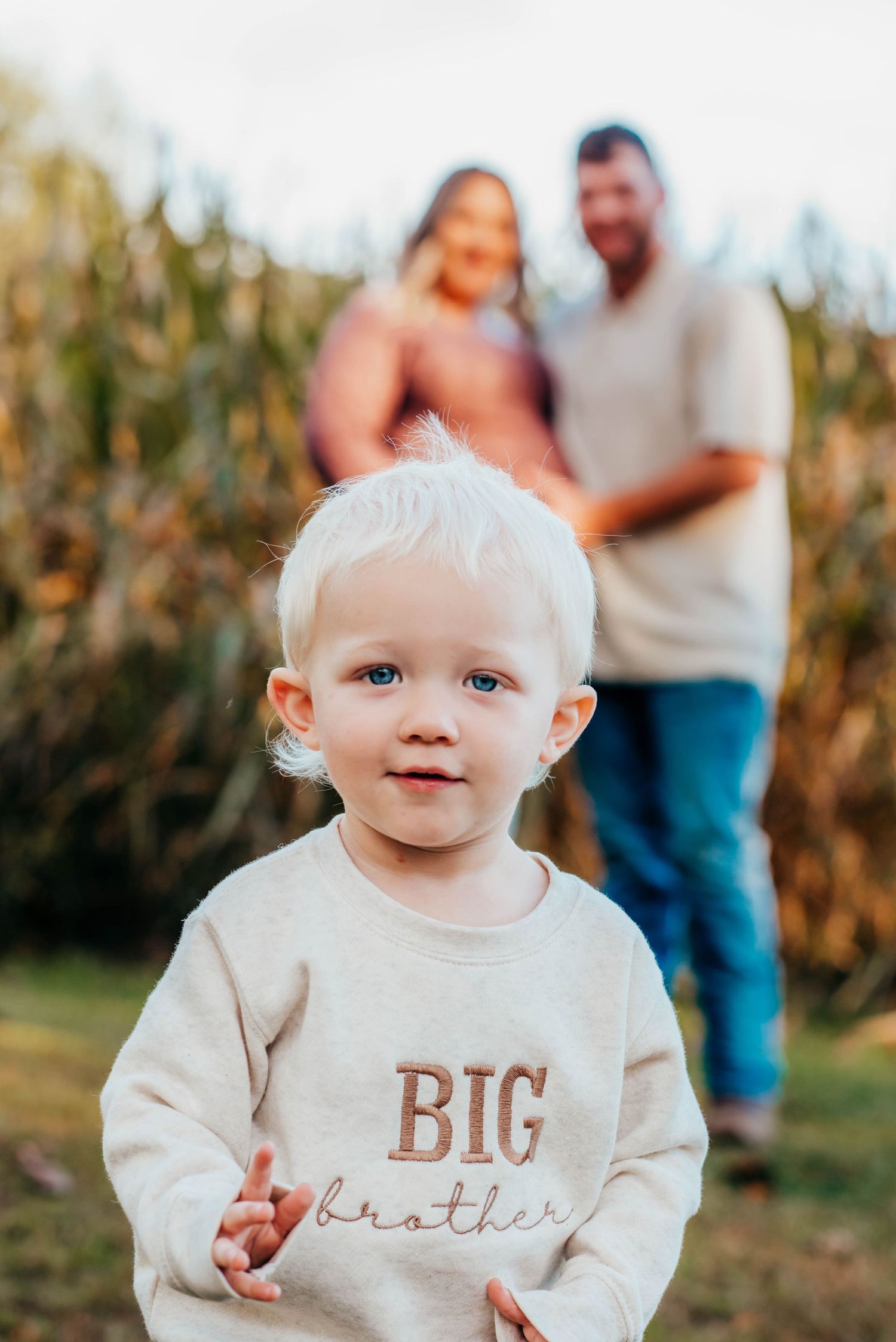 Adorable baby in white sweater smiles while sitting in autumn cornfield with family in background.