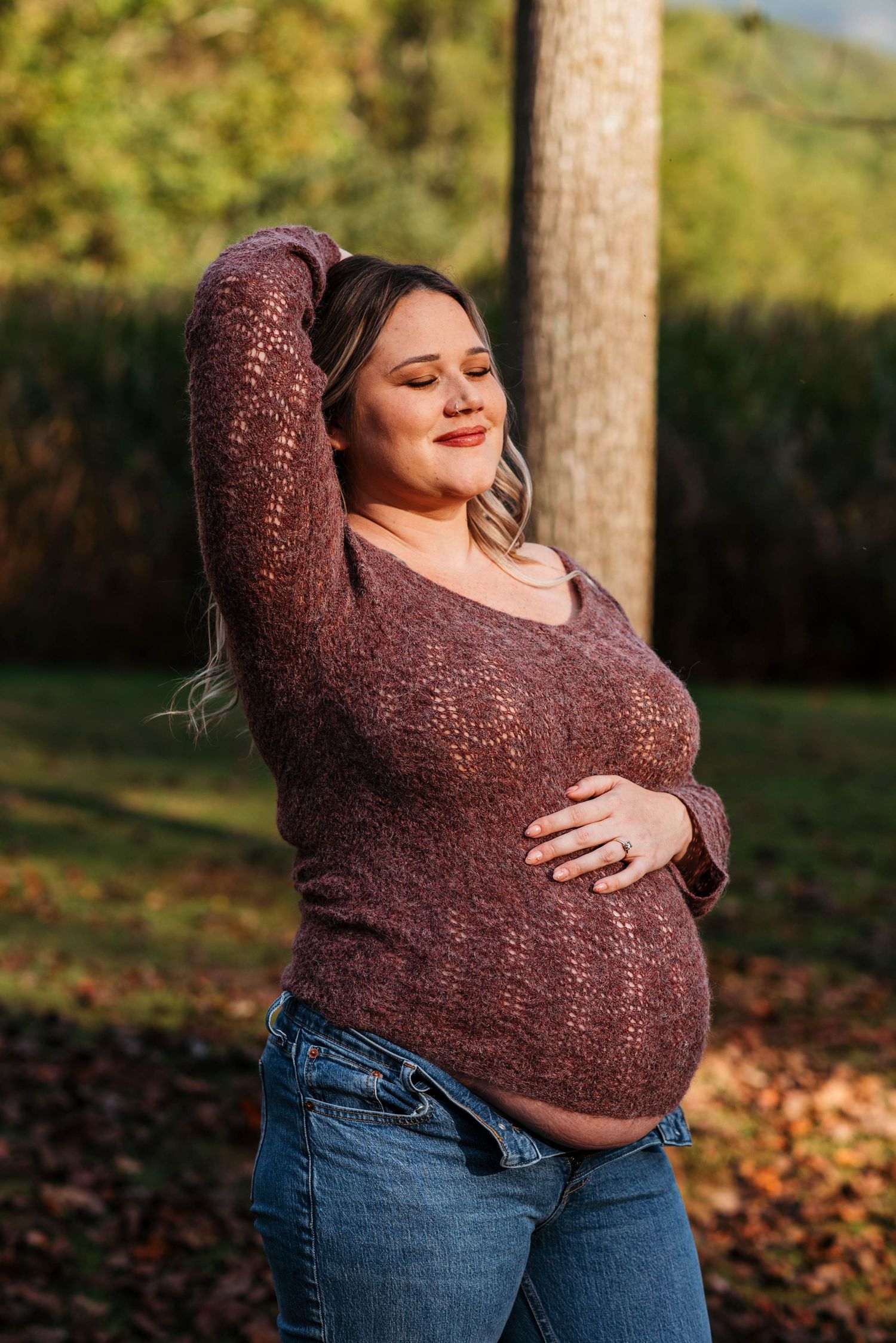 A person in jeans and a brown sweater embraces themselves outdoors on an autumn day with fallen leaves.