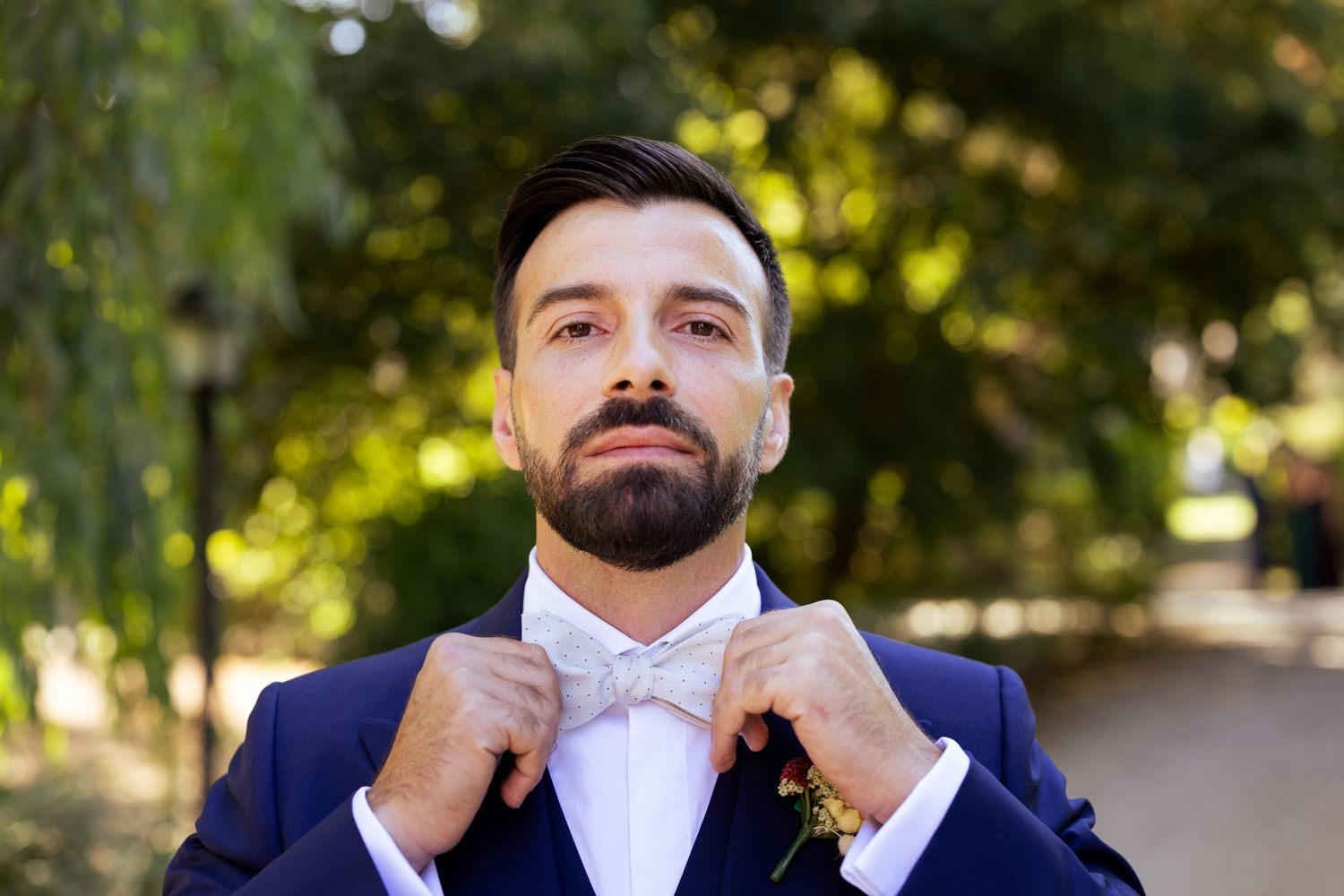 A groom adjusts his bowtie while wearing a navy blue suit and white dress shirt in an outdoor setting.