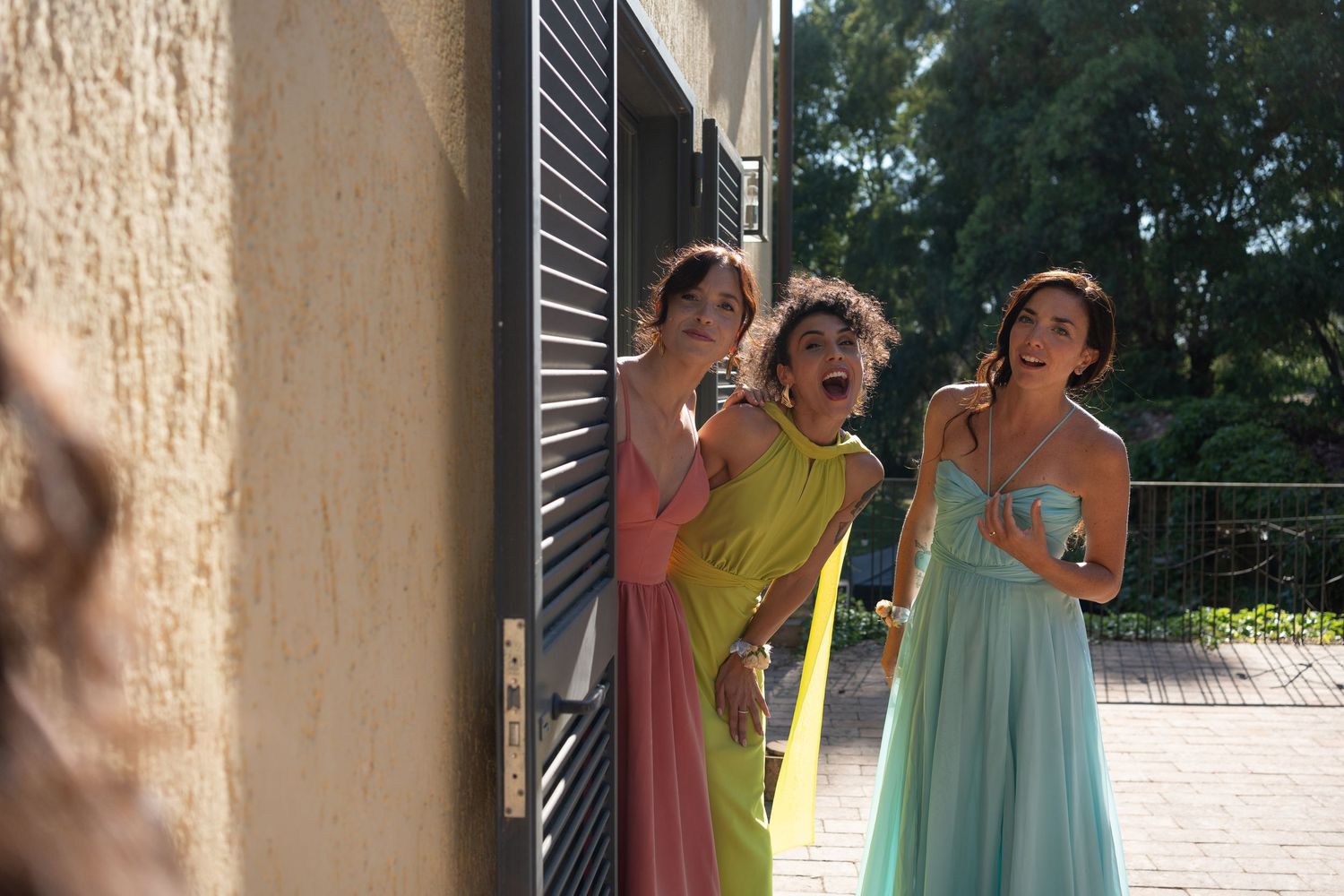Bridesmaids in pastel dresses chatting outdoors near a brick wall.