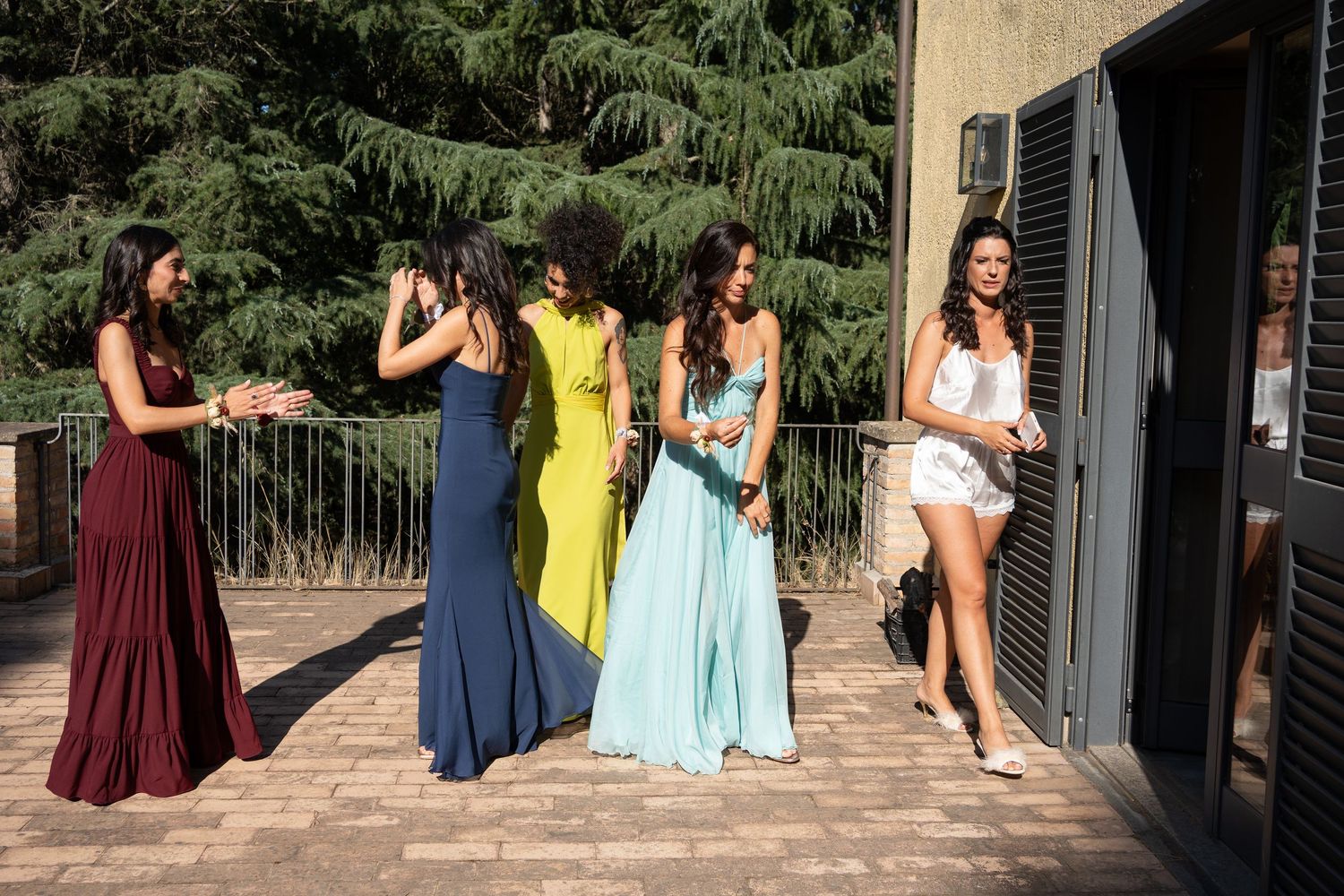 Wedding party in colorful dresses standing on a terrace with trees in the background.