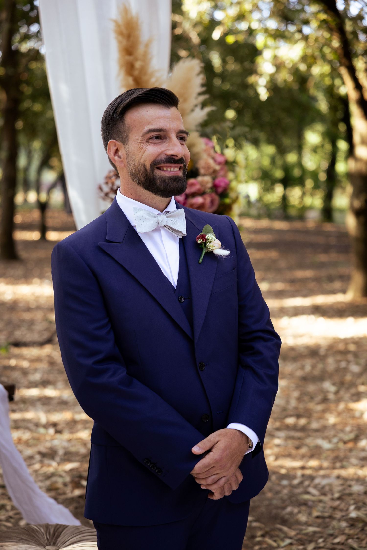 A groom in a navy suit smiling during an outdoor wedding ceremony.