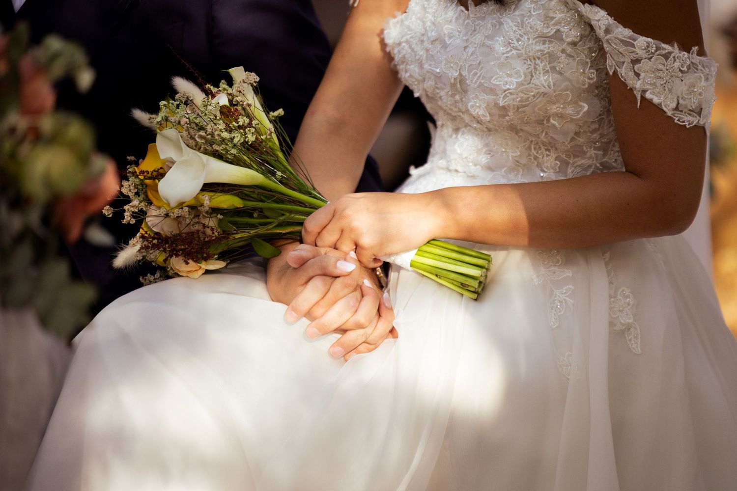 Close-up detail of hands holding a bridal bouquet with white lace dress fabric visible.