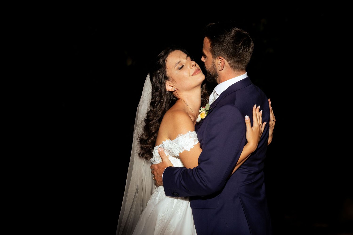 Couple sharing a romantic dance moment in formal wedding attire against dark background.