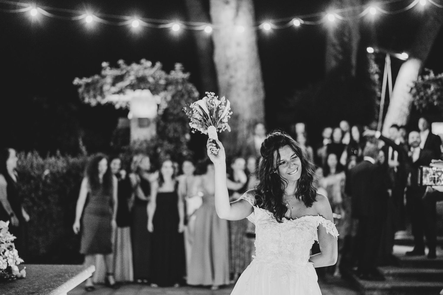 A bride in an off-shoulder white dress raises her bouquet at a nighttime outdoor reception under string lights.