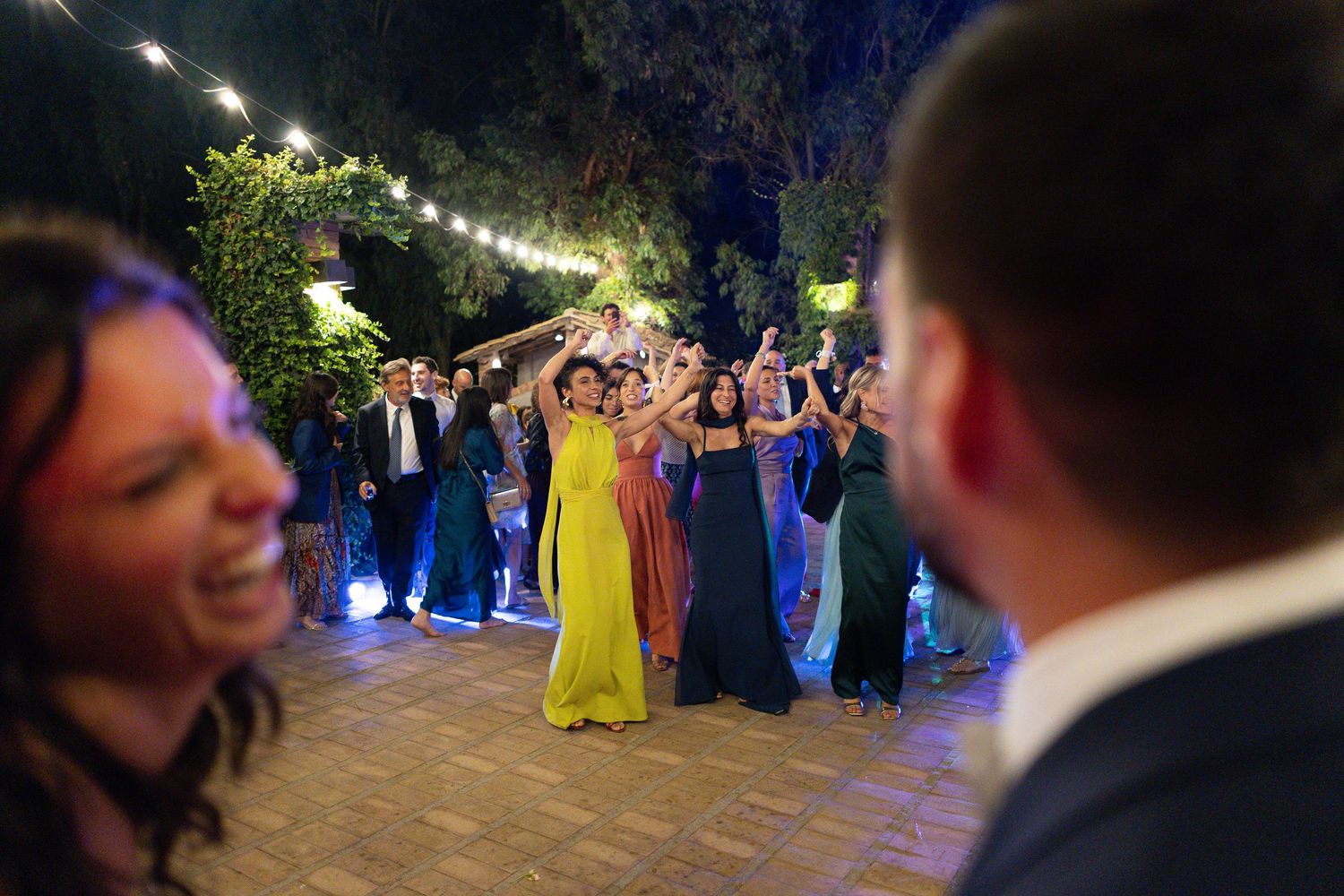 Wedding guests gather on an outdoor patio for nighttime dancing and celebration under twinkling lights.