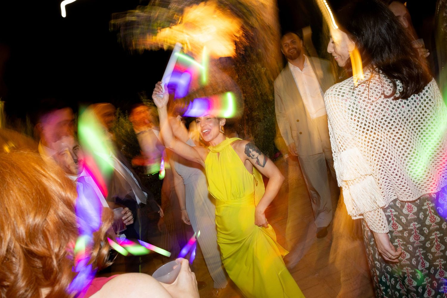 Blurred motion shot of people dancing at a party with colorful neon lights and a person in yellow dress visible.