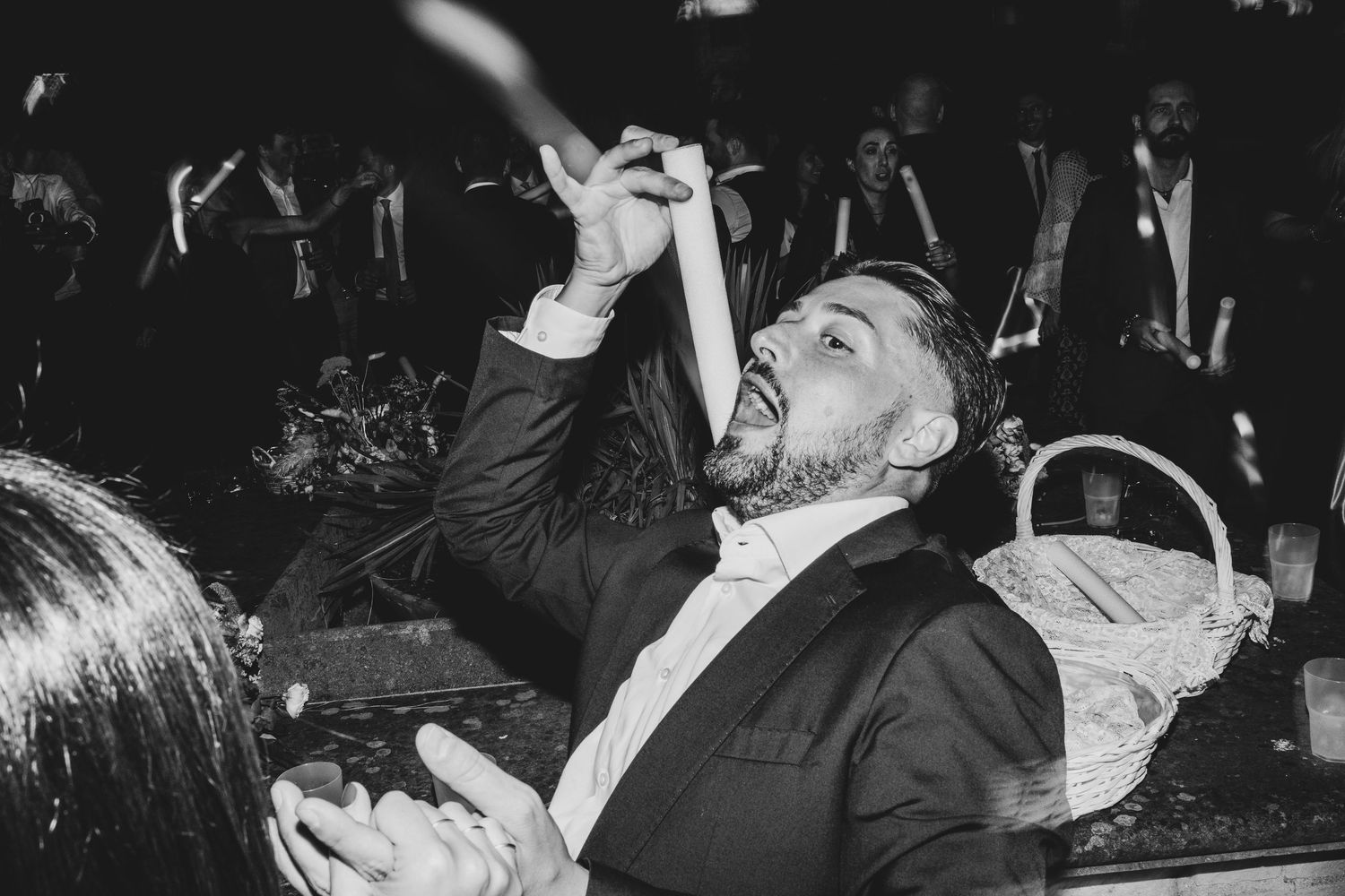 Wedding guests dance and celebrate joyfully at a lively reception party with dramatic black and white lighting.