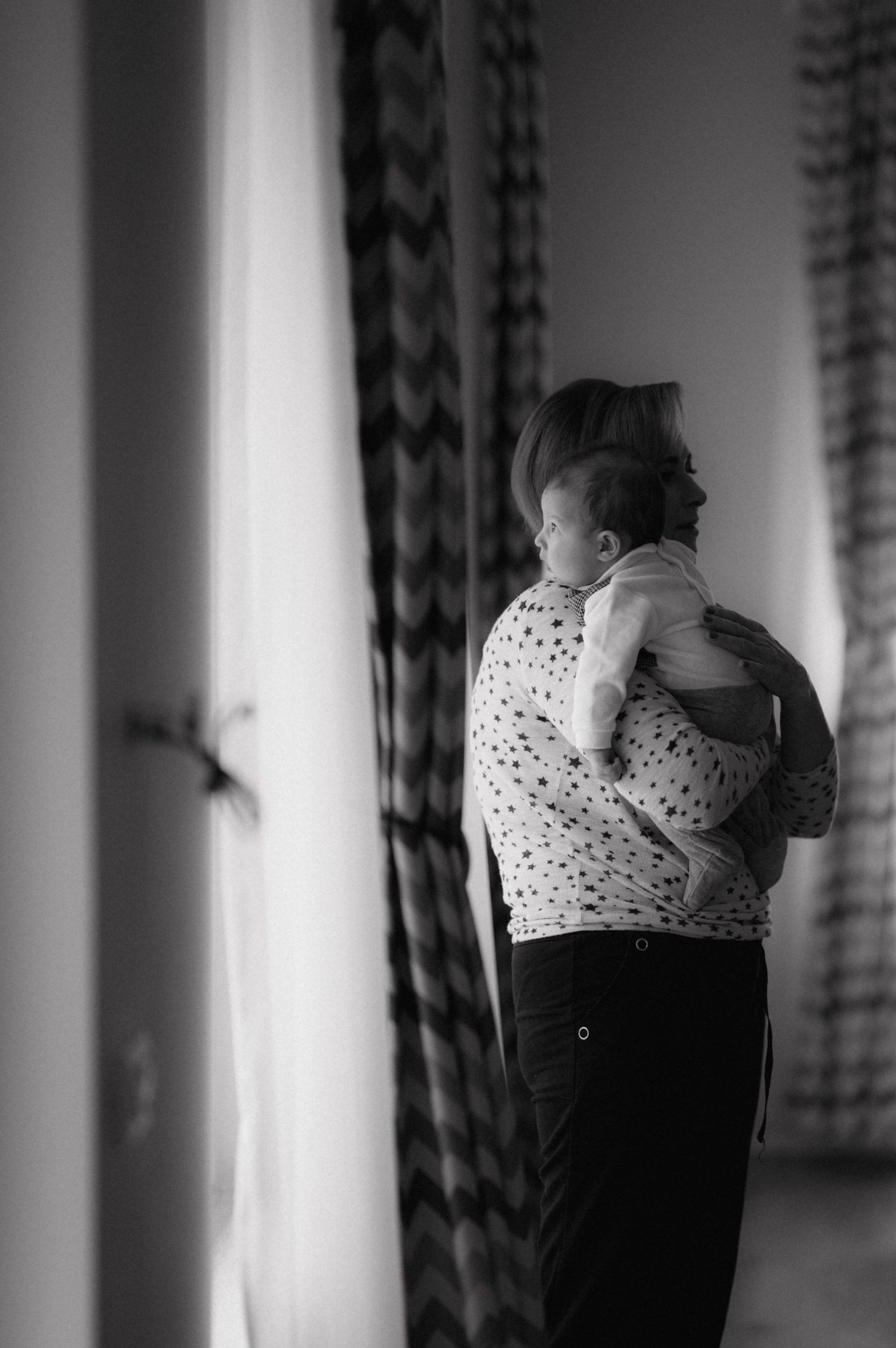 A tender moment captured in black and white as a child looks out a window with patterned curtains.